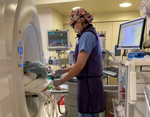 An anesthesiologist working in the CT suite, showing how the CT scanner impedes access to the patient and can make viewing the monitors and ventilator challenging.